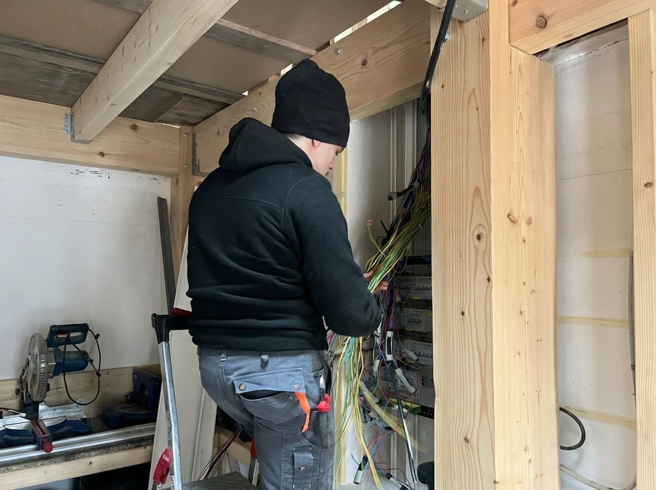 Électricien de dos, bonnet noir, installe un tableau électrique dans une construction neuve en bois